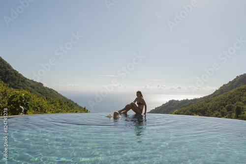 Young smiling mother sit on the edge of infinity swimming pool with sea viev and look at her child swimming in the pool, summer family vibes at vacation