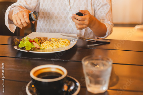 Woman having breakfast with hands putting black pepper on scrambled eggs.