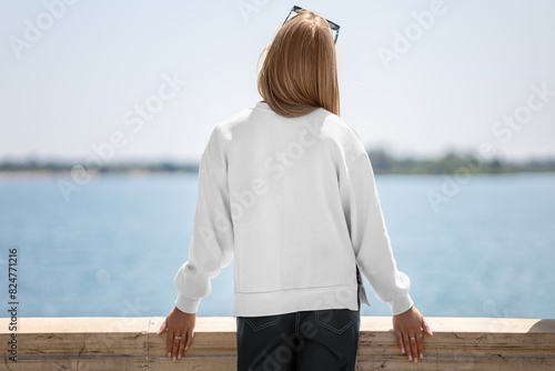 White sweatshirt template with side slits on girl, back view, lady on quay, blurred river background.