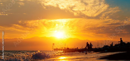 Fototapeta Naklejka Na Ścianę i Meble -  Panoramic summer background with silhouettes of people by the sea.  Beach-goers at the seashore during golden hour in summer in Antalya, Turkey.