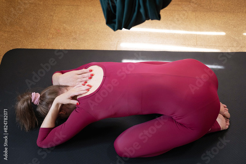 A young woman in a burgundy yoga suit is lying in the Utthita-balasana pose on a practice mat. practicing asana in a yoga studio.