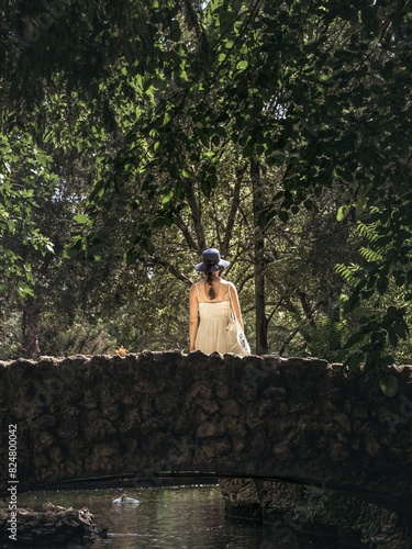 A young woman girl in a dress and hat from behind back standing on a rock stone bridge in a forest park during summer