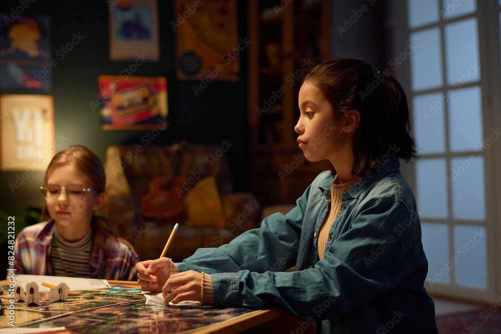 Fototapeta premium Side view of calm girl with pencil in hand looking attentively at one of players of tabletop leisure game against her friend