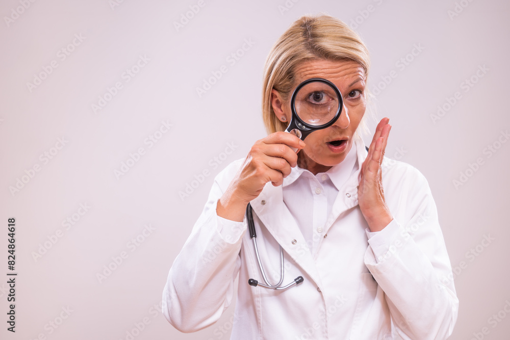 Portrait of shocked mature female doctor  with loupe on gray background.