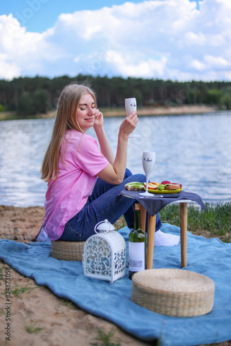 women with glass of wine or limonade making picnic outside. Positive model sitting close to lake, hugging, eating fruits and cheese