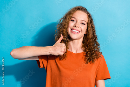 Portrait photo of youngster cheerful girl with beautiful curly red hair wearing orange t shirt showing like symbol isolated on blue color background photo