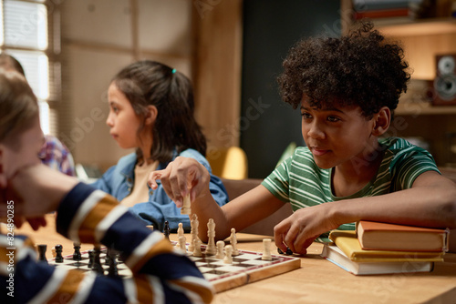 Youthful African American boy looking at checkmate sitting in front of him and holding white pawn or other figurine over chess board photo