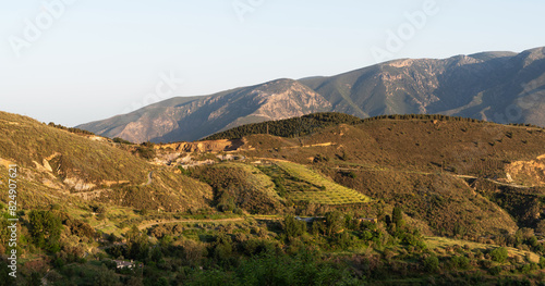 a vast landscape with layered mountain ranges, the sunlight casting soft shadows and highlights on the ridges, with lush greenery in the foreground under a clear sky.