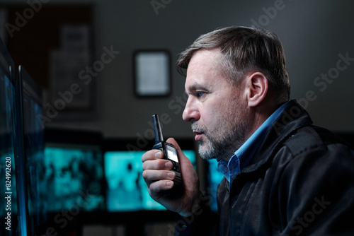 Side view of confident mature male guard looking at computer screen and reporting situation to colleagues on walkie-talkie
