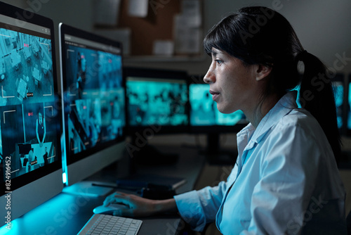 Side view of young serious Hispanic female security guard sitting by desk in control room and observing cctv video on computer screen