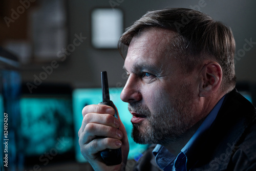 Confident mature man with walkie-talkie in hand observing situation on computer screen in control room and reporting it on transceiver photo
