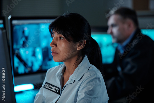 Young serious brunette woman in uniform of security guard sitting in front of computer monitor in surveillance room and looking at screen