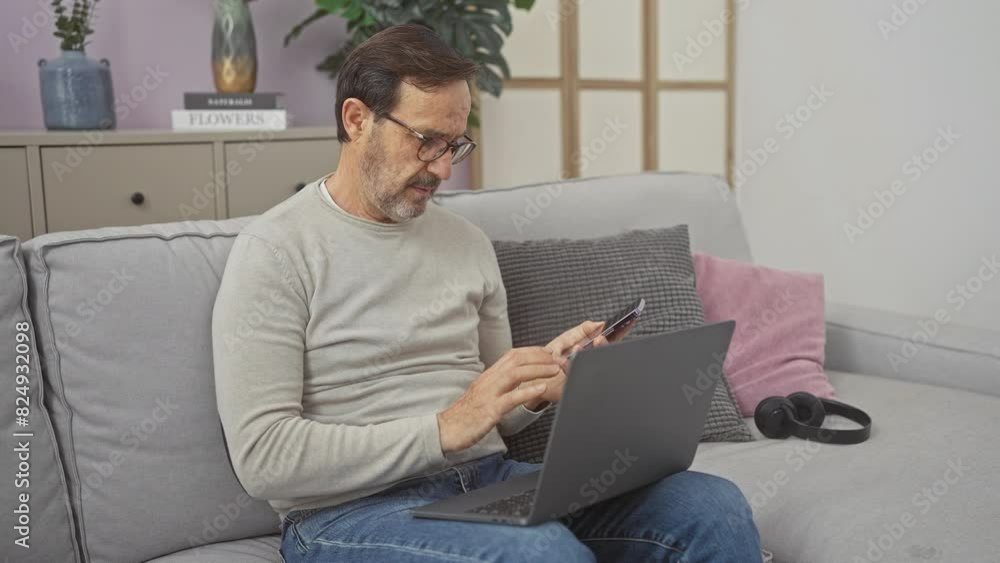 A mature man multitasks with a smartphone and laptop on a couch, indicating productivity in a casual home setting. A mature man multitasks with a smartphone and laptop on a couch, indicating productivity in a casual home setting.