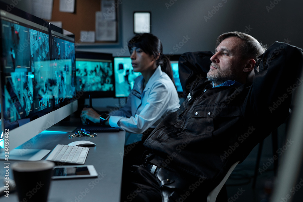 Mature male officer in black uniform keeping hands behind head while sitting in armchair in front of computer screen with cctv video Mature male officer in black uniform keeping hands behind head while sitting in armchair in front of computer screen with cctv video
