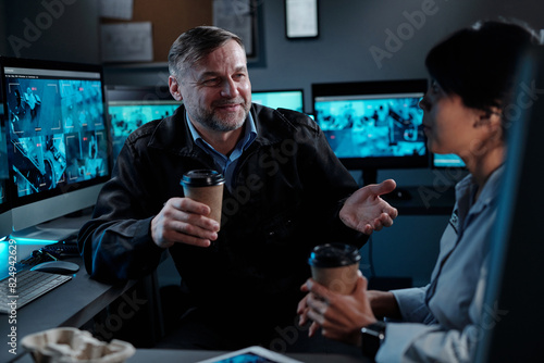 Smiling mature male security guard with cup of coffee looking at young female colleague during discussion of latest news at break photo
