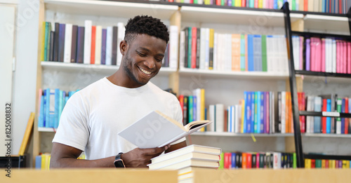 Cheerful student studying in library, holding book, sitting by bookshelf, smiling, focused on academic research.