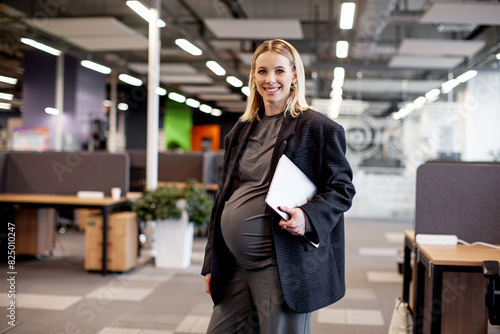 Confident pregnant businesswoman standing in a modern office, holding a tablet and smiling, showcasing balance between career and motherhood in a professional environment