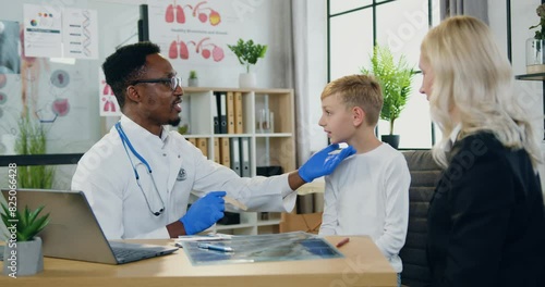 African american male doctor examining her teen son's throat with wooden spatula during their visit to clinic woman watching how professional photo