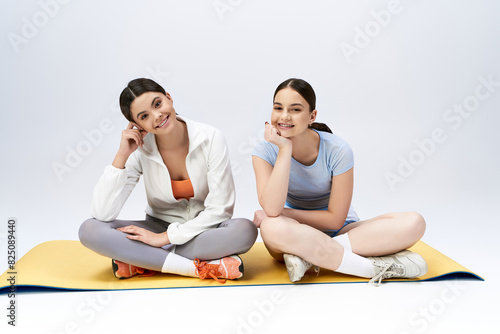 Two teenage girls, pretty and brunette, sitting on a yoga mat in a studio. They are smiling and posing for a picture.