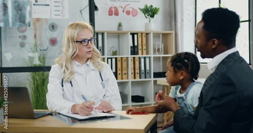 Woman doctor which writing information into medical card during father visiting medical center with his cute small daughter and telling girl's disease symptoms. Medicine concept. photo