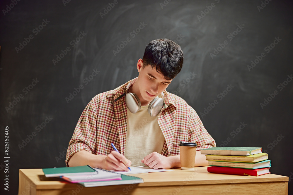 Classic Study Session. Young man sitting at wooden desk, focused on writing notes. Headphones on neck and textbooks beside. Preparing for exams. Education, studying, homework, youth, lifestyle concept Classic Study Session. Young man sitting at wooden desk, focused on writing notes. Headphones on neck and textbooks beside. Preparing for exams. Education, studying, homework, youth, lifestyle concept