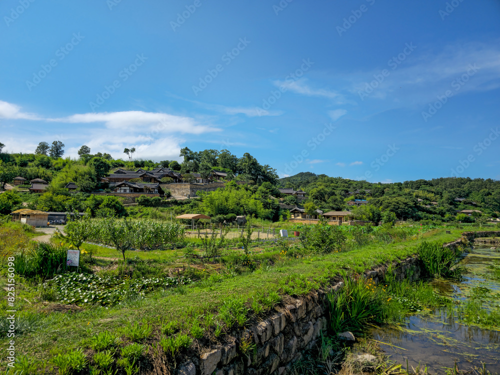 A lush green hillside with a small village in the distance. The sky is clear and blue, and the sun is shining brightly A lush green hillside with a small village in the distance. The sky is clear and blue, and the sun is shining brightly