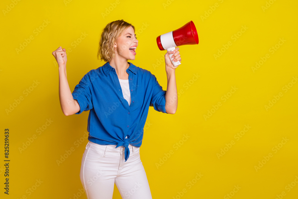 Portrait of pretty young lady loudspeaker raise fist empty space wear blue shirt isolated on yellow color background Portrait of pretty young lady loudspeaker raise fist empty space wear blue shirt isolated on yellow color background