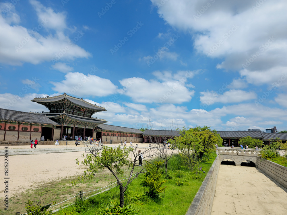 A large building with a courtyard in front of it. The courtyard is filled with people and trees. The sky is blue with some clouds A large building with a courtyard in front of it. The courtyard is filled with people and trees. The sky is blue with some clouds