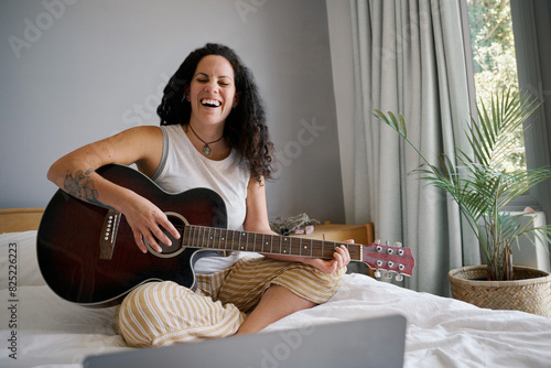 Smiling woman playing guitar on bed with laptop photo