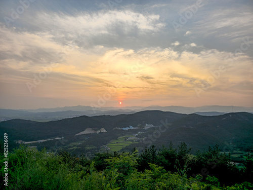 The sky is filled with clouds and the sun is setting. The mountains in the background are covered in trees © oybekostanov