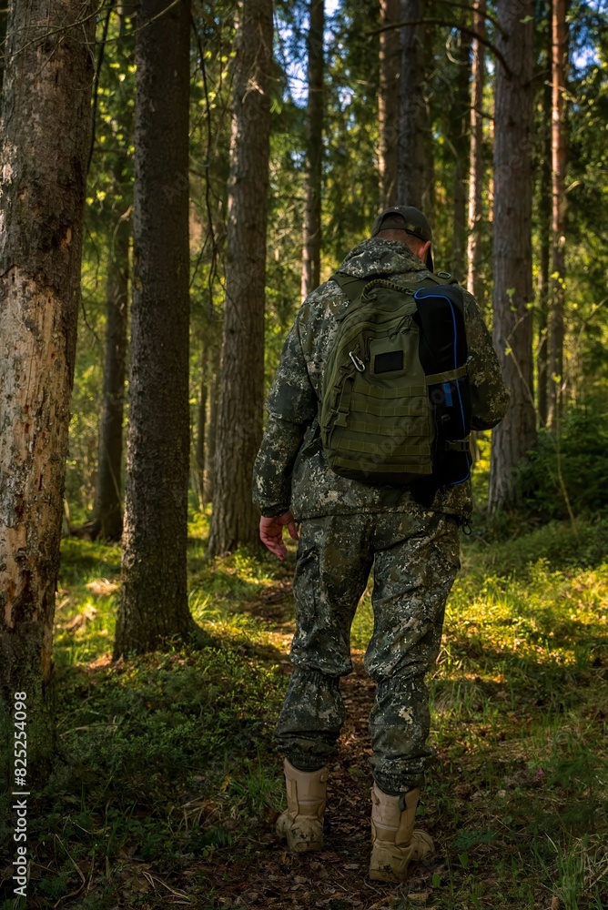 View from the back of a brutal man in a military uniform standing in a forest. A ranger with a backpack on a forest path.