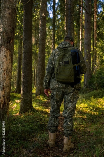 View from the back of a brutal man in a military uniform standing in a forest. A ranger with a backpack on a forest path.