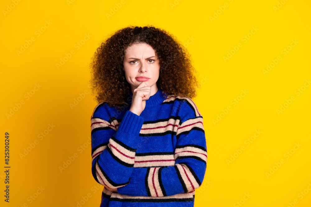 Portrait of minded puzzled gloomy girl with wavy hairstyle wear knit pullover holding hand on chin isolated on yellow color background Portrait of minded puzzled gloomy girl with wavy hairstyle wear knit pullover holding hand on chin isolated on yellow color background