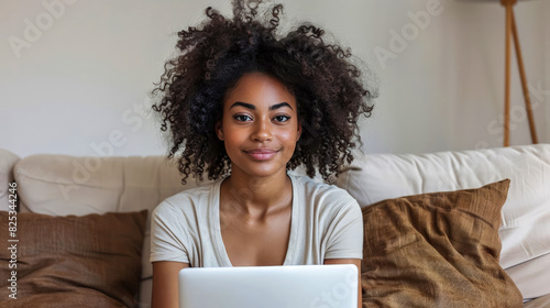 Black Woman Sitting on Couch Using Laptop at Home During Day. Young girl with curly hair is sitting on beige couch, using laptop, and smiling at camera in cozy living room. Freelancer creates content photo