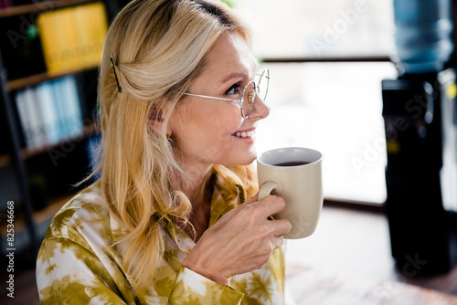 Photo portrait of lovely mature woman hold drink coffee look empty space dressed stylish shirt office day light home interior workspace