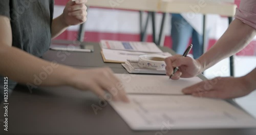Medium shot, profile, young, red-haired woman, poll worker at voting center, checks identification for male voter. He signs in and she starts handing him a ballot. Voting booths and US flag behind