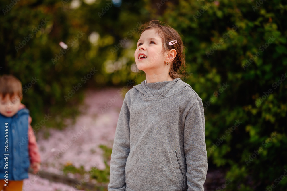 Young girl and her little brother playing outdoors on a spring day, surrounded by fallen cherry blossom petals and lush greenery. Young girl and her little brother playing outdoors on a spring day, surrounded by fallen cherry blossom petals and lush greenery.