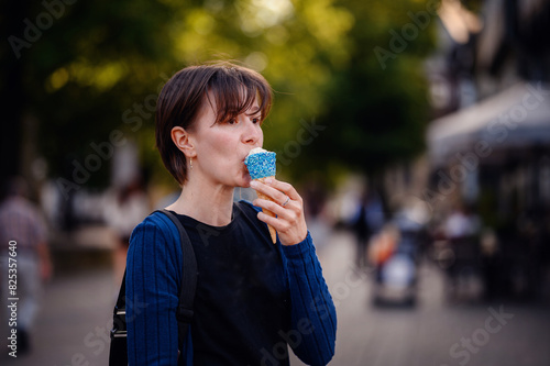 A woman with short hair enjoys an ice cream cone decorated with blue sprinkles on a sunny day in town. The background shows a lively street scene with blurred people and greenery.