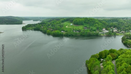 An aerial panorama of a lakeside village with houses nestled among green hills and trees, reflecting in the water. photo