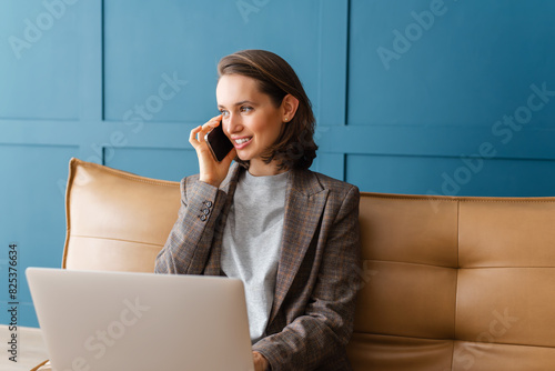Beautiful woman with brown hair sitting on the sofa, working on laptop and talking on mobile phone.