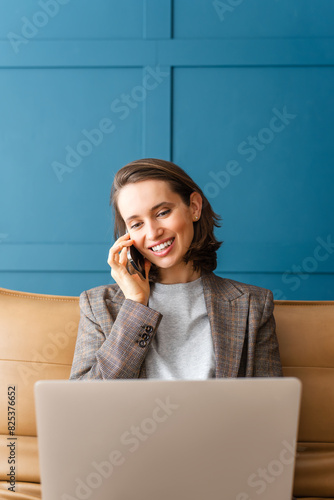 Beautiful woman with brown hair sitting on the sofa, working on laptop and talking on mobile phone.