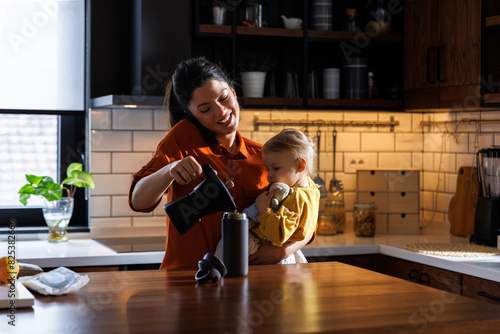Mother using phone and pouring coffee while carrying daughter photo
