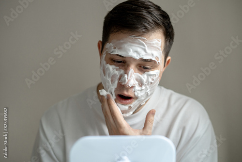 Young man applying cleaning foam on his face in front of mirror photo