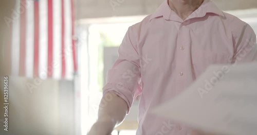 Over the shoulder, mid shot, young Asian man checks in at polling station with female poll worker using tablet computer with I voted sticker on it. Flag and voting booths behind