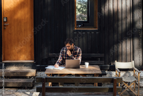 Handsome Man Sitting in Front of Mountain Cabin and Working on Laptop photo