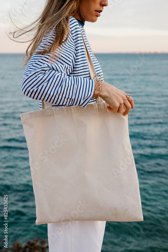 A young woman standing by the ocean, holding a plain white tote bag
