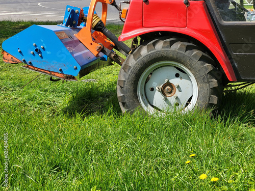 Billede på lærred A red small tractor lawn mower mows a city meadow with dandelions on a bright sp