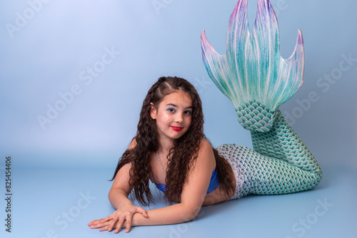 A pretty child girl in a mermaid costume lie isolated on light-blue background