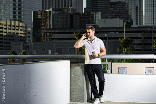 Portrait of handsome male model chatting on phone outdoor. Stylish man talking on phone dressed in polo. Fashion male posing on the street background. Business phone conversation. Urban style. photo