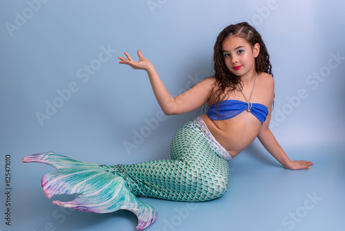 A beautiful elegant girl in a mermaid costume lies on a blue background with her palm raised up.